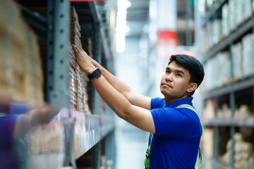 A man in a blue shirt is reaching for a box on a shelf in a warehouse. He is wearing a wristwatch...
