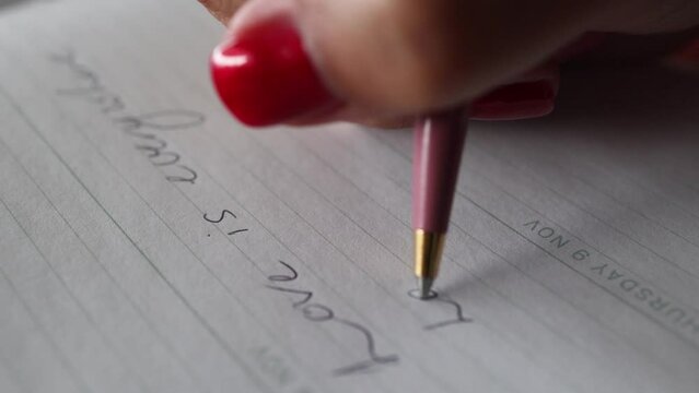 A lovestruck girl writes the phrase LOVE IS with a pink ballpoint pen in a notebook on white paper.
