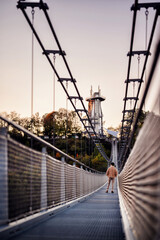 An individual in casual attire takes a solitary walk across a suspension bridge, with the twilight sky casting a soft backdrop to the autumn scenery