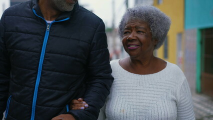 African American senior lady strolling in street with the help of her caretaker middle aged adult...