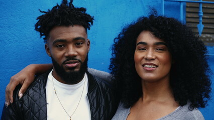 Young latin couple of African descent smiling at camera standing on blue wall in urban setting....