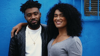 Young latin couple of African descent smiling at camera standing on blue wall in urban setting....
