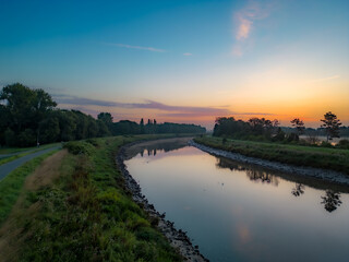This tranquil image captures the first light of dawn reflecting on the still waters of a canal, with the pastel hues of the morning sky mirrored on its surface. A pathway runs alongside the canal