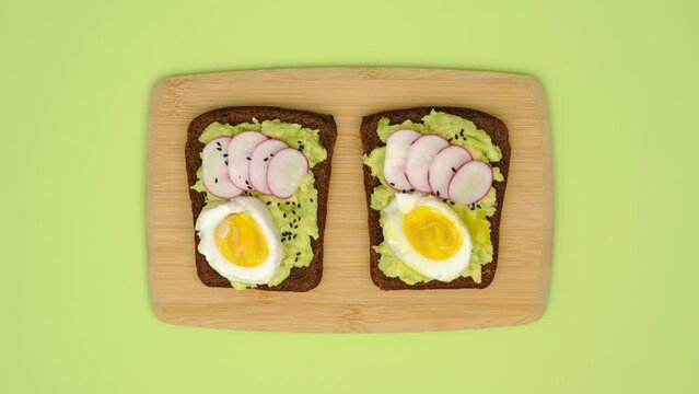 Making and eating rye bread sandwiches with avocado, egg, radish, greenery and black cumin seeds. Stop motion animation 4K video. Healthy eating concept.