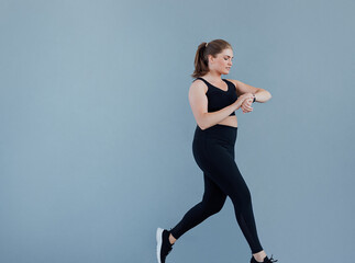 Side view of young plus-size female checking smart watch while jogging at a grey wall. Woman in black sportswear looking at her smartwatch while running.
