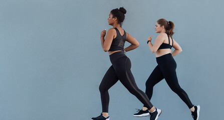 Full length of two plus size women running synchronized at a grey wall. Two women in fitness wear...