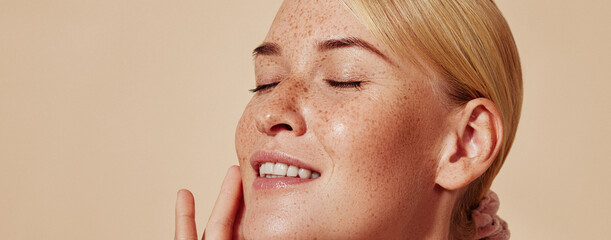 Side view of smiling blond woman with freckles touching her face. Close-up studio shot of young positive female with closed eyes against pastel background.