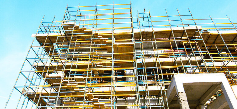 A building under construction with scaffolding and yellow and blue safety rails. The building is tall and has a lot of scaffolding