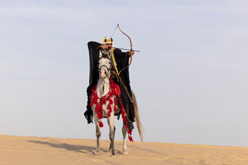 Saudi man in traditional clothing shooting an arrow in the desert while siting on a white horse