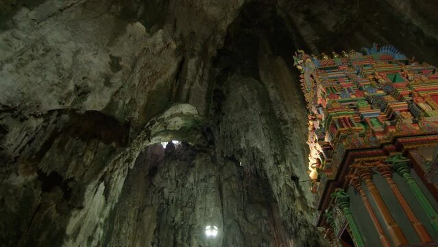 limestone walls and ceiling of the Batu Caves, punctuated by natural light from openings above, alongside a vibrant section of Hindu temple architecture, richly adorned with carvings and statues