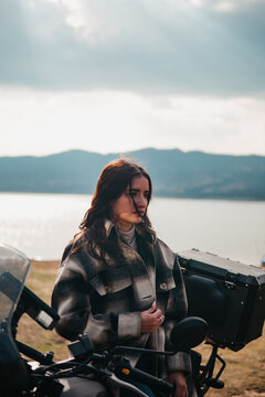 A contemplative Latina woman stands by her motorcycle, looking off into the distance by a serene lakeside with mountains in the backdrop