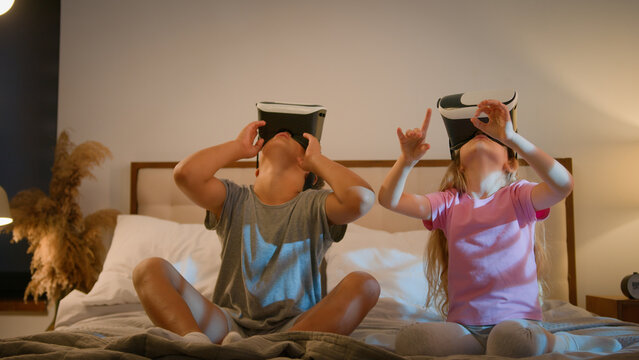 Funny little amazed caucasian children kids boy girl brother sister playing video game using digital virtual reality headset play online metaverse 3d with VR glasses in bedroom innovative technology - Powered by Adobe