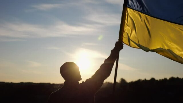 Male ukrainian army soldier lifting blue-yellow banner in honor of the victory against russian aggression at sunset. Young man in camouflage uniform raising a waving flag of Ukraine at countryside