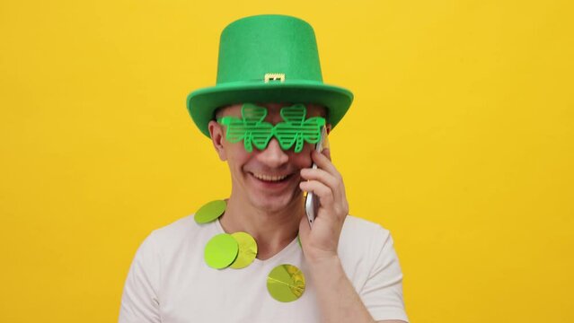 Smiling cheerful positive man wearing green leprechaun party hat isolated over yellow background talking on mobile phone congratulating with holiday expressing happiness - Powered by Adobe