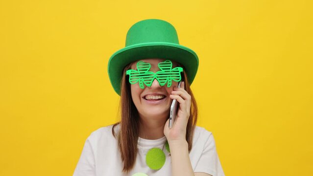 Cheerful St. Patrick's Day leprechaun model female in green hat and clover  glasses isolated over yellow background talking with friend on mobile phone laughing happily