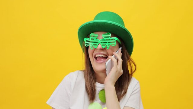 Happy overjoyed woman wearing leprechaun party green hat and clover glasses isolated over yellow background talking on mobile phone with happy facial expression laughing