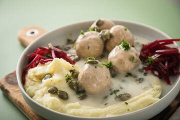 Königsberger Klopse mit Kapern, Kartoffelpüree und Rote Beete Salat in Schale mit  Holzbrett, Silber Besteck und Serviette auf Hintergrund grün