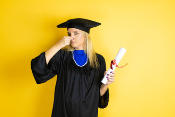 Beautiful blonde young woman wearing graduation cap and ceremony robe smelling something stinky and disgusting, intolerable smell, holding breath with fingers on nose