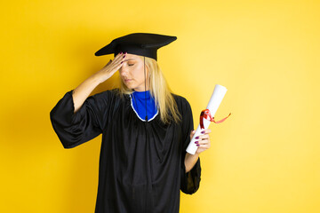 Beautiful blonde young woman wearing graduation cap and ceremony robe putting one hand on her head...