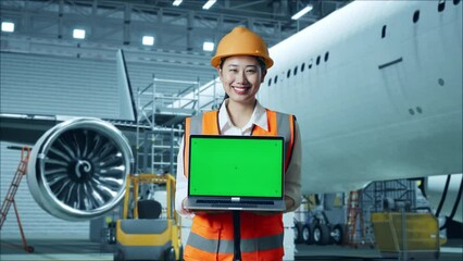 Asian Female Engineer With Safety Helmet Standing With Aircraft In The Hangar. Smiling And Showing Green Screen Laptop To The Camera While Aircraft Maintenance