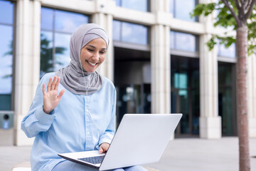 Cheerful Muslim lady wearing a hijab engaging in a video chat, waving to the webcam with a joyful smile outside.