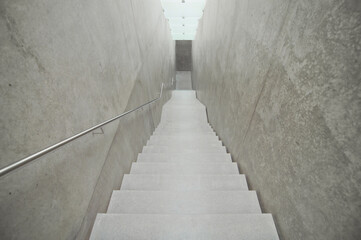 Empty narrow downwards staircase. Concrete walls and illuminated ceiling made of satin glass....