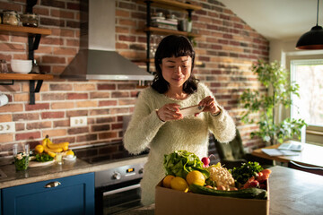 Content Asian woman photographing vegetables in sunlit kitchen