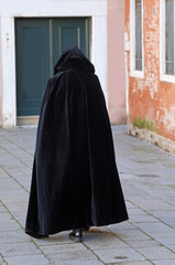figure in a hood walks through a narrow city alley wearing a worn black tabard