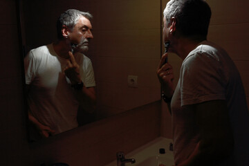 Man with gray hair and a defect on his hand shaving in front of the mirror with foam and razor. Low...