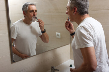 Man with gray hair and a defect on his hand shaving in front of the mirror with foam and razor....