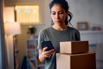 Latin American woman with delivered packages texting on mobile phone at home.