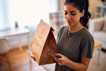 Hispanic woman texting on cell phone after receiving home delivery in paper bag. - obrazy, fototapety, plakaty
