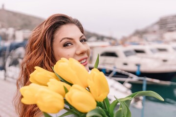 Woman holds yellow tulips in harbor with boats docked in the background., overcast day, yellow...