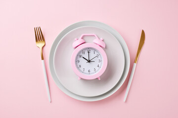 Eating schedule adherence: timely eating habits. Top view shot of a pink alarm clock, cutlery,...