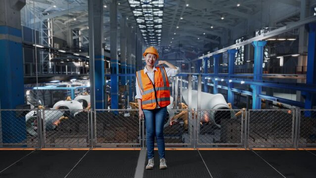 Full Body Of Asian Female Engineer With Safety Helmet Standing In Factory Manufacture Of Wind Turbines. Smiling To Camera And Making Call Me Gesture While Robotic Arm Working
