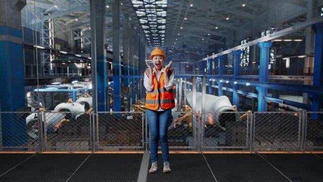 Full Body Of Asian Female Engineer With Safety Helmet Standing In Factory Manufacture Of Wind Turbines. Smiling To Camera And Saying Wow While Robotic Arm Working
