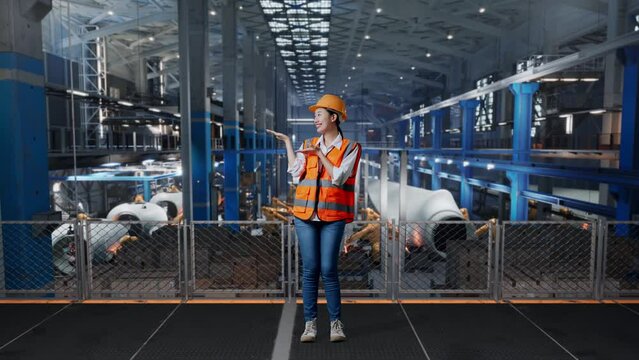 Full Body Of Asian Female Engineer With Safety Helmet Standing In Factory Manufacture Of Wind Turbines. Smiling And Pointing To Side Recommends About Something While Robotic Arm Working