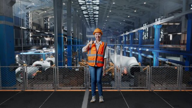 Full Body Of Asian Female Engineer With Safety Helmet Standing In Factory Manufacture Of Wind Turbines. Smiling And Showing Thumbs Up Gesture To The Camera While Robotic Arm Working