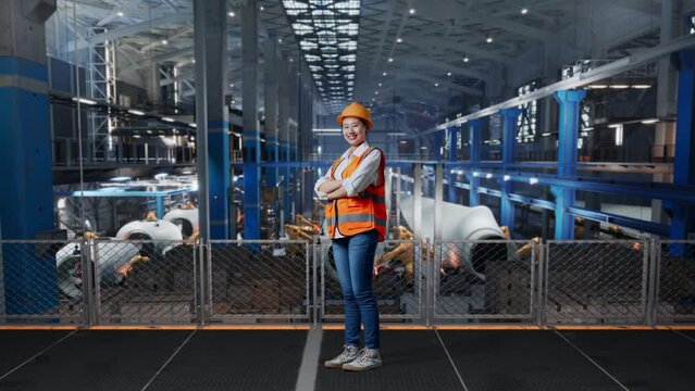 Full Body Of Asian Female Engineer With Safety Helmet Standing In Factory Manufacture Of Wind Turbines. Crossing His Arms And Smiling To Camera While Robotic Arm Working