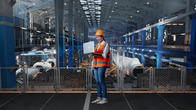 Full Body Side View Of Asian Female Engineer With Safety Helmet Looking At A Laptop And Looking Around While Standing In Factory Manufacture Of Wind Turbines