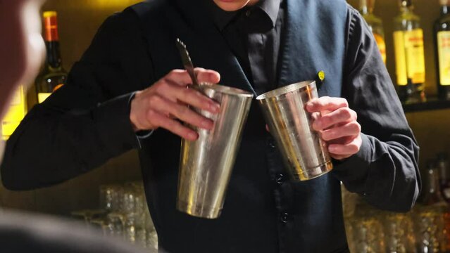 Guy works as bartender by barstand mixing cocktail in shaker. Barman pours drink from cup to shaker to mix cocktail for guests