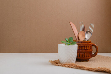 ceramic mug with kitchen cuttlery and small pot with plant on table with space for text 