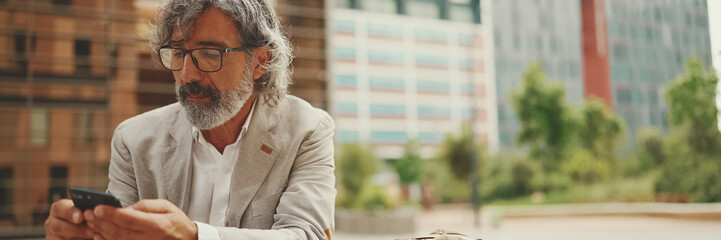 Mature businessman with beard in eyeglasses wearing gray jacket is using cell phone, Panorama....