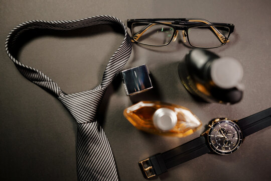 Banner of glasses, tie, and a bottle of cologne. The bottle of cologne is on a table next to a watch in an elegant setting