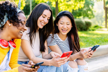 Three young women using mobile phones sitting together at city park in a sunny day. Technology lifestyle and social media concept.