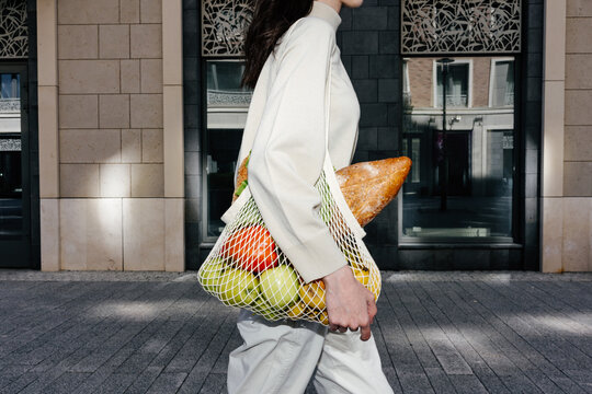 Crop woman walking along street with mesh bag of groceries 