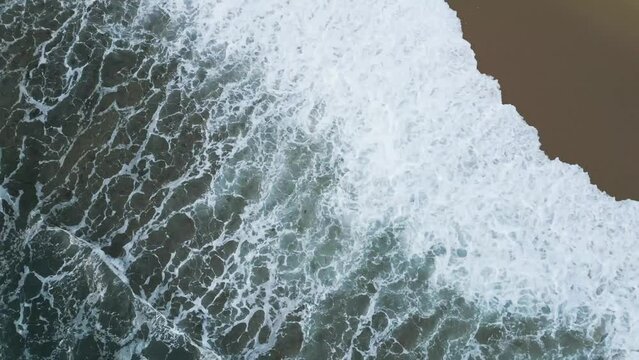 Drone bird's eye view over the deep blue ocean waves and beach. Beautiful waves roll onto the sandy shore at sunset. Water texture, 4K