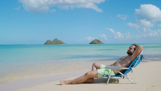 Barechested man with beard in beach shorts relaxing, napping in a folding chair, holding arm under his head, scenic azure waters of ocean touching his feet. High quality 4k footage