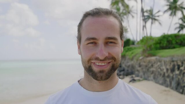 Attractive fair-haired unshaved guy in white T-shirt standing on the scenic beach of Hawaii island, smiling at camera, showing white straight teeth. High quality 4k footage