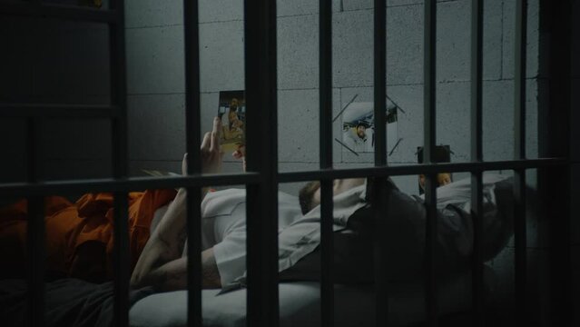 Male prisoner lies on bed in prison cell, looks at pictures of family. Inmate or criminal serves imprisonment term for crime in jail. View through metal bars. Detention center or correctional facility